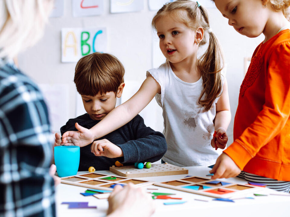 Two beautiful little girls standing at table with pencils in bright classroom. Little boy sitting, playing board games.